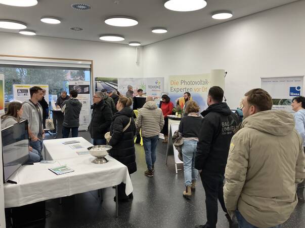 In einem hellen Raum findet eine Messe oder Ausstellung statt, bei der sich mehrere Personen an Ständen zu Themen rund um Photovoltaik informieren.