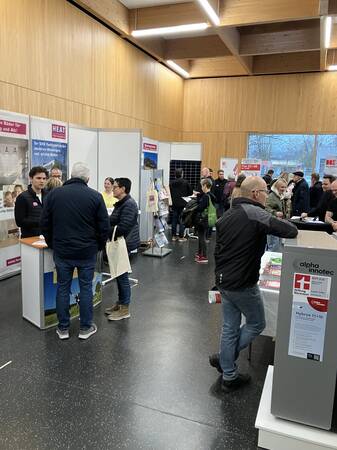 In einem Raum mit Holzverkleidung finden Gespräche an mehreren Messeständen statt, an denen sich Personen informieren und beraten lassen.