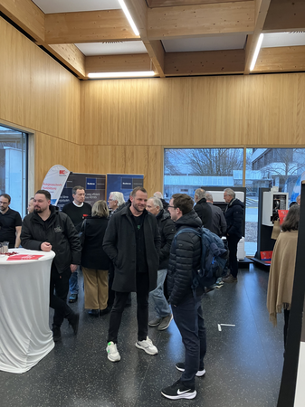 In einem modernen Raum mit großen Fenstern sind mehrere Menschen bei einer Veranstaltung oder Messe in Gespräche vertieft, während Infostände im Hintergrund zu sehen sind.