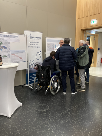 Mehrere Personen stehen vor Infotafeln, die über die zukünftige Wasser- und Wärmeversorgung informieren, und eine Person im Rollstuhl beteiligt sich im Gespräch.