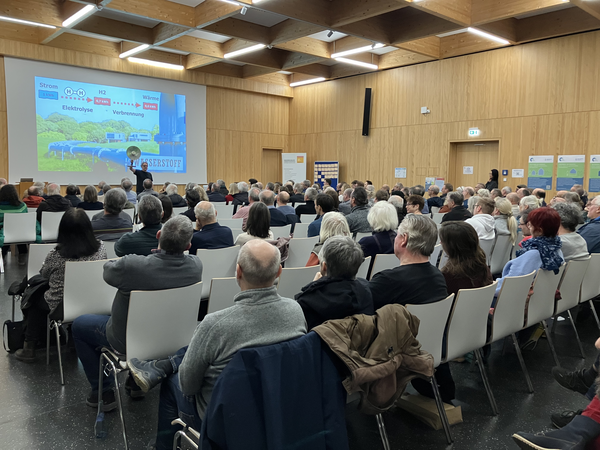 Eine große Gruppe von Menschen sitzt in einem modernen Konferenzraum und hört einem Vortrag zu, während eine Präsentation über Wärmepumpen auf einer großen Leinwand gezeigt wird.