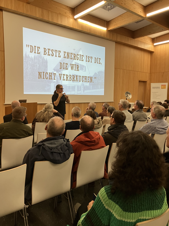 Eine Person hält einen Vortrag vor einem Publikum in einem Holzkonferenzraum; im Hintergrund ist eine große Leinwand mit dem Zitat "Die beste Energie ist die, die wir nicht verbrauchen." sichtbar.