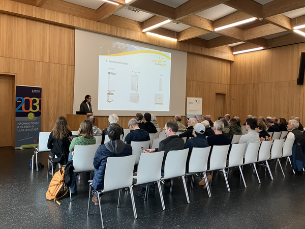 Ein Vortragender steht vor einem Publikum in einem modernen Holzsaal und spricht vor einer Leinwand, auf der eine Präsentation gezeigt wird.