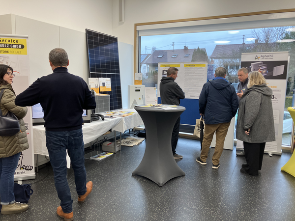 Mehrere Menschen unterhalten sich in einer Ausstellungshalle mit Informationsständen und Roll-ups, die Solartechnologie und Batterien thematisieren.