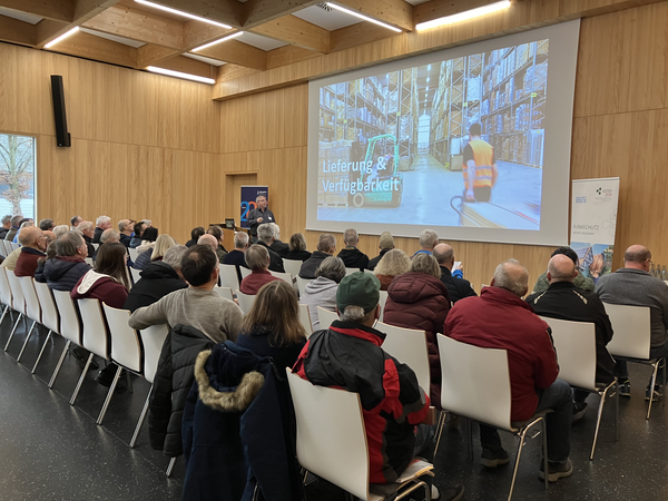 Eine Gruppe von Menschen sitzt in einem Saal und hört einem Vortrag zu, während auf einer großen Leinwand ein Bild mit dem Titel "Lieferung & Verfügbarkeit" angezeigt wird.