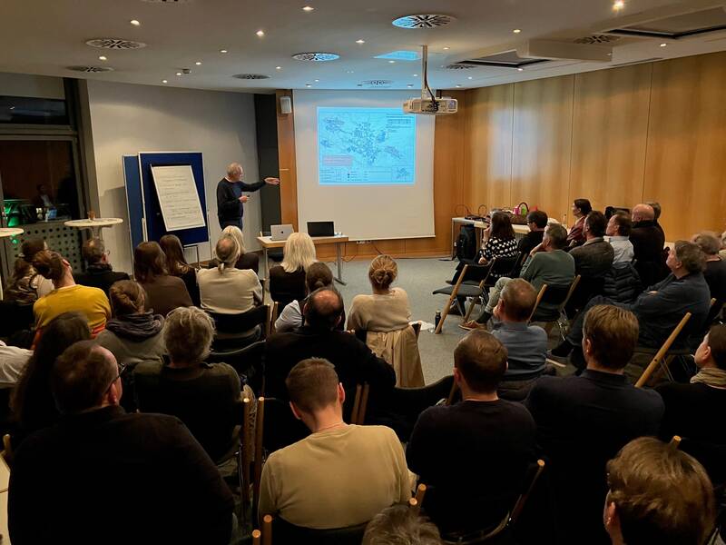 Eine Person hält eine Präsentation vor einem sitzenden Publikum in einem Konferenzraum mit Projektion und Flipchart.